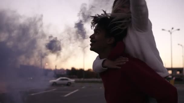 Gros plan de deux jeunes couples fous tourbillonnant avec des bombes fumigènes colorées dans les mains sur la zone de stationnement vide à l'extérieur. Des amis sur le dos. Émotions, mode de vie heureux, rire — Video