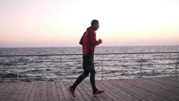 Filmato laterale di un giovane sportivo che fa jogging, riscaldandosi prima dell'allenamento principale di boxe. Esercizi mattutini vicino al mare, pugni in aria — Video Stock