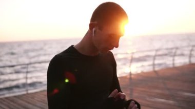 Beyaz adam boksör için egzersiz deniz kenarında ahşap zemin üzerinde dururken elleri siyah renkli bandaj ile kaydırma hızlandırılmış görüntüleri. Kablosuz kulaklık ile müzik dinlemek