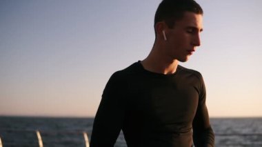 Genç atletik erkek üzerinde beyaz kulaklıklar koyarak siyah tişörtlü adam ve onun smartphone üzerinde müzik geçiş. Müzik açık denizin içinde açık havada yaz dinle