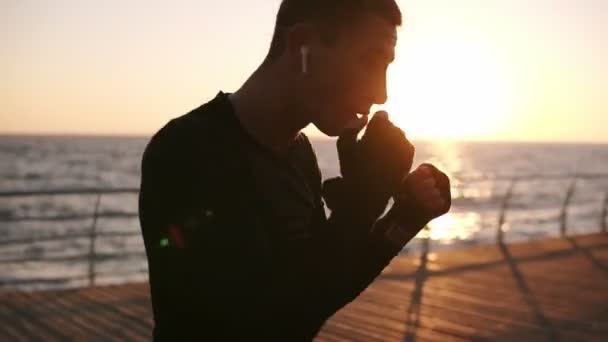 Jonge mannelijke bokser in zwarte shirt en rode sneakers tijdens een training verwerken op de promenade aan de voorzijde van de zee in de vroege ochtend. Man boksen met onzichtbare tegenstander, ponsen - witte koptelefoon dragen — Stockvideo