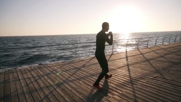 Lunghezza completa di un giovane atleta in camicia nera e scarpe da ginnastica rosse durante il processo di allenamento sul lungomare di fronte al mare la mattina presto. Uomo boxe con avversario invisibile, punzonatura indossando nero — Video Stock