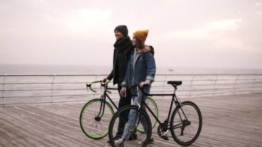 Gülümseyen birkaç güzel genç hipsters onların bisiklet ile birlikte kucaklayan yürüyüş deniz sonbahar gün yakın. Sarı şapkalı bir şeye işaret eden genç kız. Ahşap güverte tarafından gündüz yürüyüş