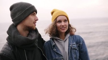 Birlikte ayakta ve deniz kenarında sarılma hipster genç mutlu bir çift. Genç insanlar sıcak giysiler, soğuk havalarda şapka giyiyor. Gülümseyen arıyorsunuz