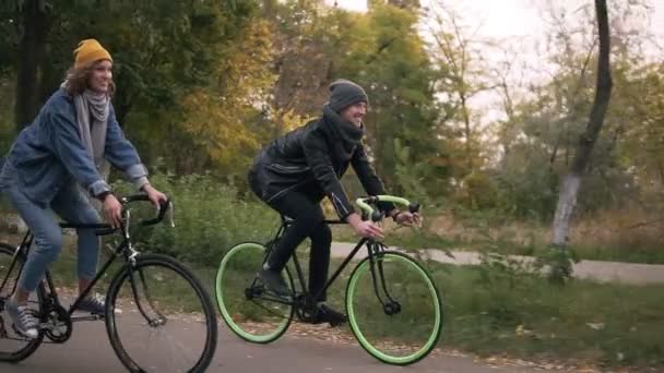 Para młoda hipster się na rowerze po parku na rowerach trekkingowych. Dwóch młodych ludzi świetnie bawimy razem jesienią. Widok z boku — Wideo stockowe