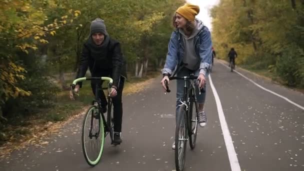 Jeunes, amis souriants ou jeune couple en chapeaux faisant du vélo sur leurs vélos de randonnée à travers le parc d'automne sur des vélos. Homme et femme faisant du vélo ensemble et parlant. Vue de face — Video