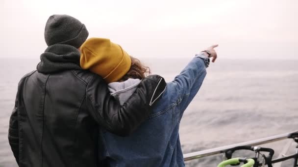 年轻人带着一个女孩站在海上木墩上拥抱, 年轻夫妇享受时间。戴黄色帽子的女孩指着在海面上的地平线上。秋天。罕见的视图 — 图库视频影像