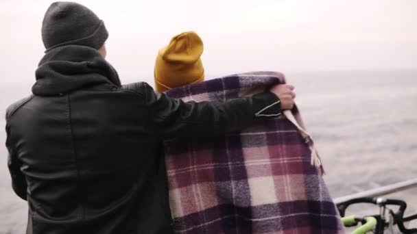 Hombre joven se puso el cuadros en los hombros de sus amigas mientras estaba de pie abrazándose en el muelle de madera de mar, pareja joven disfrutando del tiempo. chica con sombrero amarillo hablando con su novio. Otoño. Vista rara — Vídeos de Stock