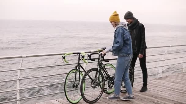 Χαμογελά δυο νέοι Χίπστερς ξεκινώντας περπατώντας μαζί με τα ποδήλατά τους κοντά στη θάλασσα, τη μέρα του φθινοπώρου. Το περπάτημα με ξύλινο deck στη διάρκεια της ημέρας. Ορίζοντα στη θάλασσα. Αργή κίνηση — Αρχείο Βίντεο