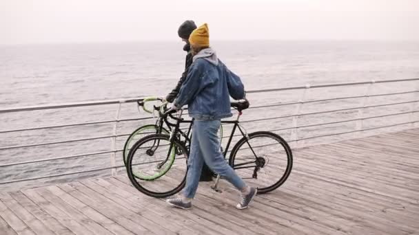 素敵な流行に敏感なカップルを残す彼らのバイクを一緒に秋の日に 海に近い受け入れを歩いてクロスバーに傾いた ウッドデッキで昼間歩いてください — ストック動画