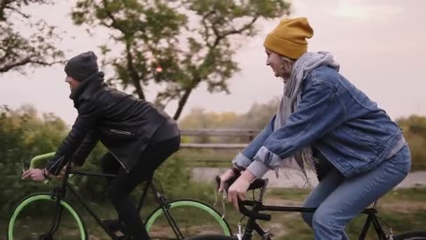 Young hipster couple enjoying cycling through park on trekking bikes. Two young people having great time together in autumn. Side view. Slow motion — Stock Video