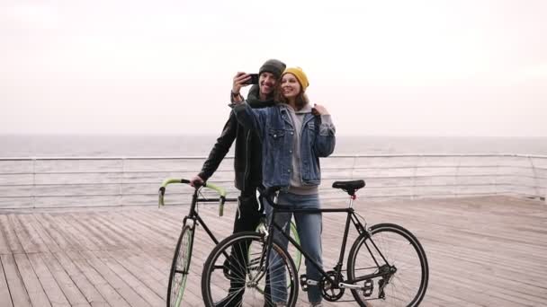 Jovem casal feliz tomando selfie por telefone celular perto da beira-mar em dia nublado. Menina de chapéu amarelo segurando o smartphone, posando. Bela selfie casal, amor e conceito de bicicleta. Comprimento total — Vídeo de Stock