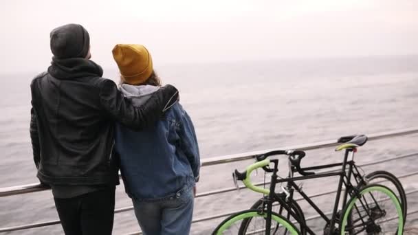 Tillbaka porträtt av unga par stående nära havet, kärlek par koncept, par med cyklar, par bära varma kläder, kille med en flicka som står på havet träpiren — Stockvideo