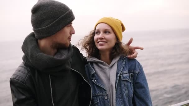 Birlikte ayakta ve deniz kenarında sarılma hipster genç mutlu bir çift. Genç insanlar sıcak giysiler, soğuk havalarda şapka giyiyor. Birbirlerine, gülümseyerek, konuşmayı arıyorsunuz. Yakın çekim — Stok video