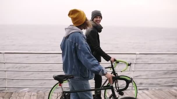 Hermosa pareja sonriente de jóvenes hipsters caminando juntos con sus bicicletas cerca del mar en el día de otoño. Caminando por la cubierta de madera en el día nublado. De cerca. Vista lateral — Vídeos de Stock