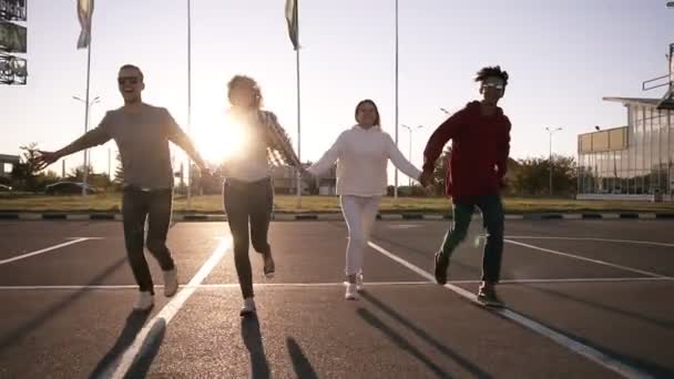 Чотири друзі біжать порожньою паркувальною зоною на відкритому повітрі - веселе щастя, молоді чоловіки і жінки біжать у вечірній сутінках і тримають разом руки — стокове відео