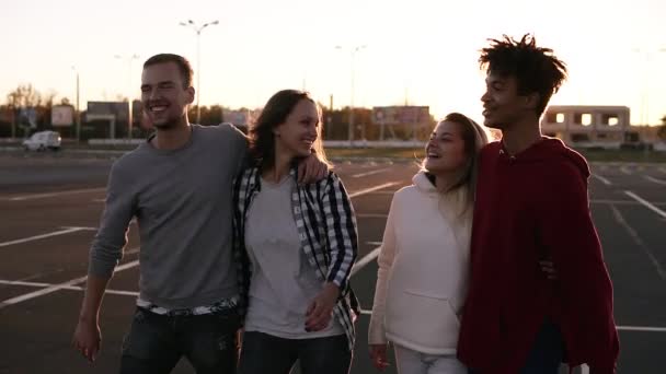 Grupo de quatro jovens amigos multirraciais rindo enquanto caminham ao ar livre. Jovens homens e mulheres desfrutando de um dia de verão na zona de estacionamento perto do shopping. Noite crepúsculo — Vídeo de Stock