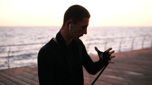 Αθλητικός άνδρας Καυκάσιος μπόξερ περιτυλίγματος χέρια με μαύρο χρωματισμένο επιδέσμους για προπόνηση ενώ στέκεται στο ξύλινο πάτωμα κοντά στη θάλασσα. Ακούστε μουσική με τα ασύρματα ακουστικά — Αρχείο Βίντεο
