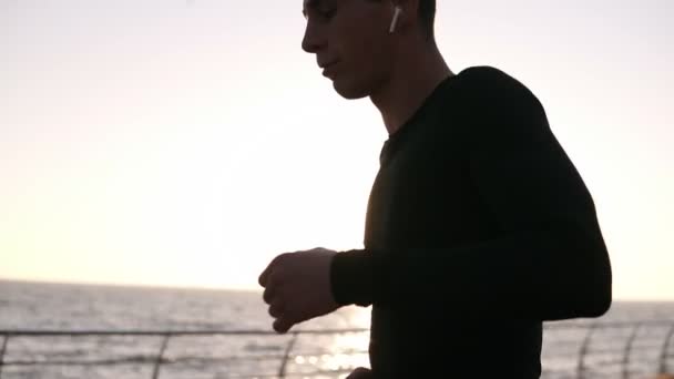 Mladý běžec zastavil, aby měl trochu odpočinout před dlouhé. On nosí sportovní černé tričko s bílým sluchátka. Brzy ráno před mořem. Slunce silně svítí na pozadí — Stock video