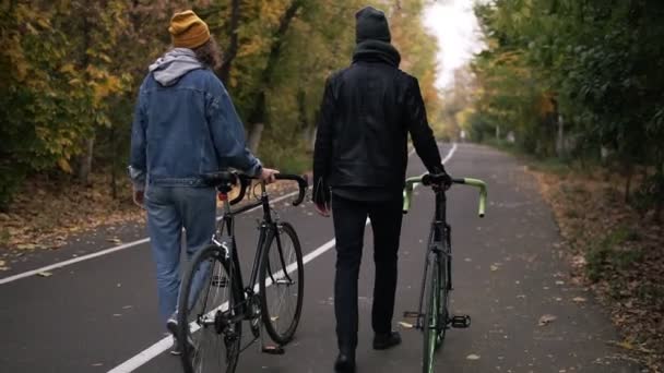 Dos amigos cercanos que tienen paseo encantador con bicicletas en el parque de la ciudad entre árboles altos verdes y amarillos, concepto de estilo de vida activo, comunicación, citas. Vista rara — Vídeos de Stock