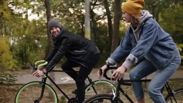 Glimlachen, jonge hipster paar genieten van fietsen door park op trekkingfietsen. Twee jonge mensen in hoeden hebben geweldige tijd samen in de herfst. Zijaanzicht. Slow motion — Stockvideo