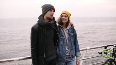 Birlikte ayakta ve deniz kenarında sarılma hipster genç mutlu bir çift. Genç insanlar sıcak giysiler, soğuk havalarda şapka giyiyor. O gülümseyen sevgilisi üzerinde seyir sarı şapkalı kız