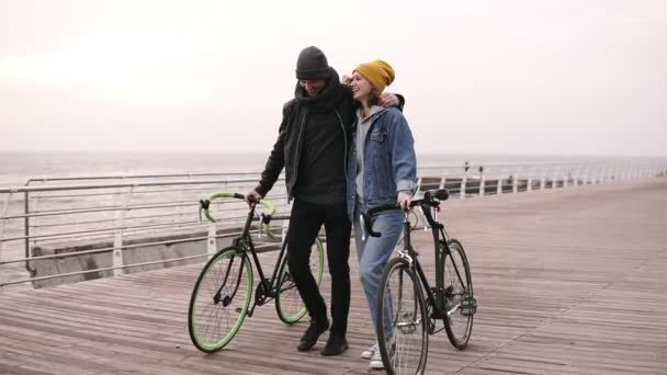 Полная длина пары молодых хипстеров, прогуливающихся вместе со своими велосипедами возле моря осенним днем. Дневная прогулка по деревянной палубе — стоковое видео
