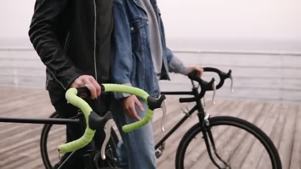 Jeune couple marchant au bord de la mer sur des vélos dans la journée nuageuse d'automne. Images de gouvernails avec les mains. Personnes, activité, concept de loisirs. Vue latérale — Video