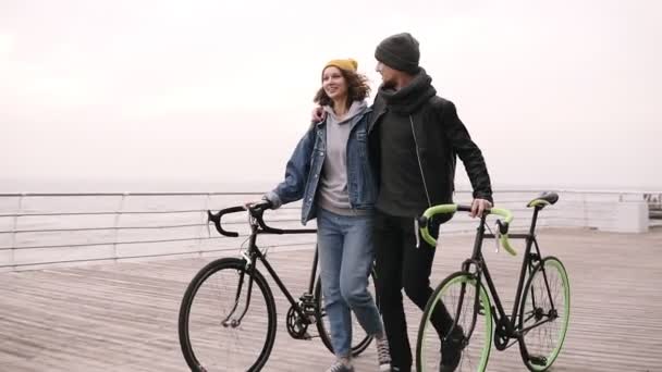 Moderna pareja hipster joven caminando junto con sus bicicletas cerca del mar en el día de otoño abrazando. Caminando por la cubierta de madera durante el día, sonriendo y hablando. Movimiento lento — Vídeo de stock