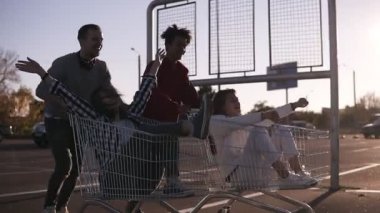 Yan görünümü bir genç arkadaşlar açık havada alışveriş arabaları üzerinde eğleniyor. Gençler çok ırklı üzerinde alışveriş arabaları park bölgesi üzerinde arkadaşlarıyla yarış. Gülüyor, eğleniyor çift