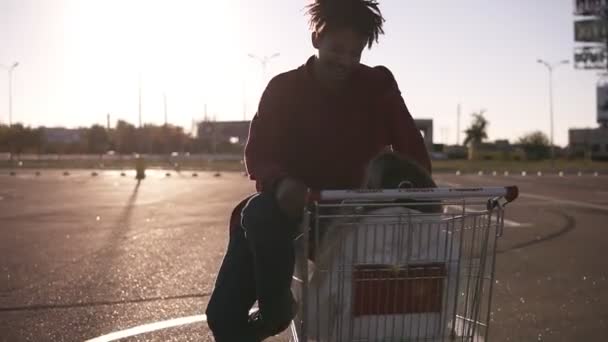 Afroamerican ung pojke med trendiga frisyr som sitter på en shopping cort på ouside parkering zonen och ser att hans flickvän. Kaukasiska flickvän sitter inne cort och prata med henne — Stockvideo