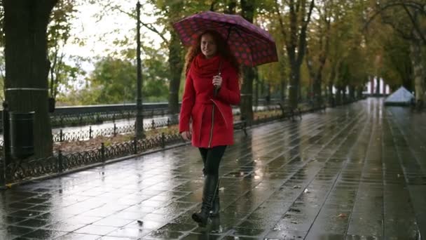 Leggenda piena di persona femminile in passeggiata rossa sotto pioggia con ombrello. Donna dai capelli rossi che indossa un cappotto rosso con ombrello passeggiando per il parco cittadino. Vista frontale — Video Stock