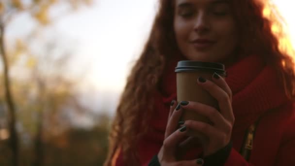 Hermosa escena de dicha de una chica de pie al aire libre en el parque y disfrutar de su café. Hermosa mujer joven con el pelo rizado rojo largo, imágenes de visión general. Chica encantadora en la luz del sol de oro que brilla desde — Vídeo de stock