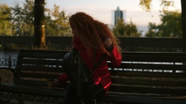 Mujer leyendo un libro en un banco del parque, tiempo libre en el parque de otoño. Preciosa mujer pelirroja con abrigo rojo sentada en un banco y sacando el libro de su bolso — Vídeos de Stock