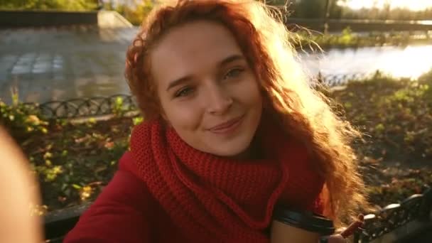 Heureuse femme caucasienne avec de longs cheveux bouclés dans un manteau chaud et foulard se promène dans le parc d'automne avec des feuilles jaunes. Sourire en regardant la caméra, cligner des yeux, poser — Video