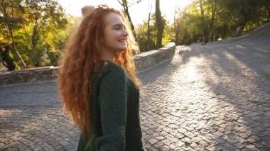 Karanlık örme kazak sevinçle renkli bir sonbahar Şehir Parkı kaldırımda yürüyen genç çekici kız. Kırmızı kıvırcık saçlı kız sonbahar yaprakları, zevk döner sevinçle kameraya gülümseyen çevresinde