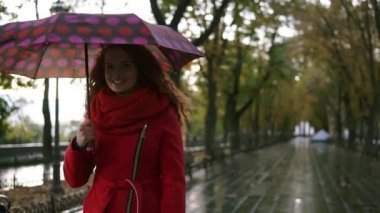 Kırmızı kafa güzel bir kadın portresi şemsiye ile yağmur altında yürümek. Şehir Parkı ile yürüyüş ve gülümseyen şemsiye ile kırmızı ceket giyen güzel, mutlu kadın
