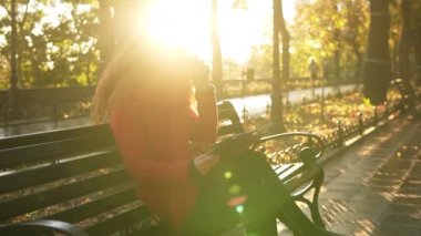Kırmızı içecekler kahve ve sonbahar hava sırasında parkta bankta oturan kadın tablet kullanma. Sosyal medya kontrol, rahatlatıcı kırmızı kafa kız. Güneş parlıyor ve arka plan üzerinde
