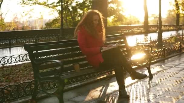 El día soleado de otoño por la tarde en el parque, la joven está leyendo un libro, la niña está sentada en el banco del parque con su bolso, hojas coloridas de otoño que cubren la acera, relajarse en el parque de la ciudad. Visión general — Vídeos de Stock