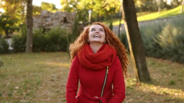 Porträtt av en glad röd huvud kvinna kasta löv runt och letar upp en höstar dag. Höstlöv faller på glad ung kvinna i stadsparken och hon virvlande runt i njutning. Leende — Stockvideo