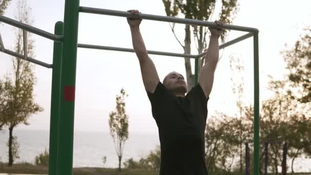 Atleta forte em roupas esportivas pretas fazendo pull-up na barra horizontal. Mans fitness com céu azul e árvores no fundo e espaço aberto em torno dele — Vídeo de Stock