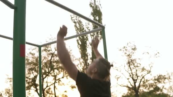 鉄棒でのプルアップを行う開始黒 sportclothes の強いアスリートは。屋外で運動する男。背景や彼の周りのオープン スペースでの木。まれなビュー — ストック動画