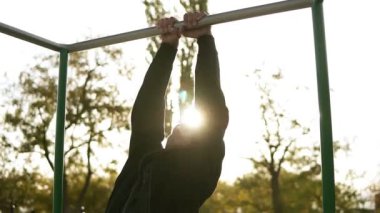 Güçlü atlet siyah sportclothes pull-up yatay çubuğu'nda yapıyor. Arka plan ve çevresindeki ağaçlar güneş parlar. Ağır çekim