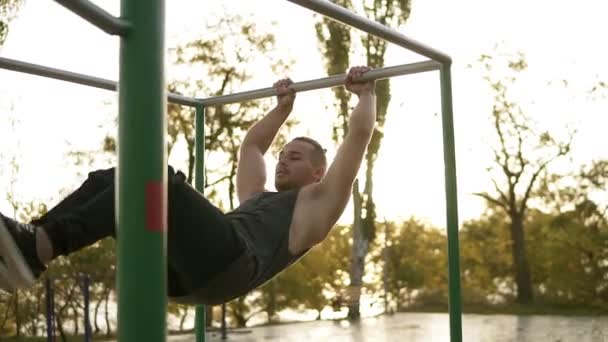 Bonito homem ter calistenia formação em barra horizontal ao ar livre. Faz acrobacias de ginástica na barra transversal no parque. Os foguetes da lente no fundo. Movimento lento — Vídeo de Stock