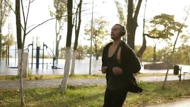 Fredlig man kör och lyssnar på musik genom hörlurar. Jogging i morgon grön park i svart casual kläder. Slow motion — Stockvideo