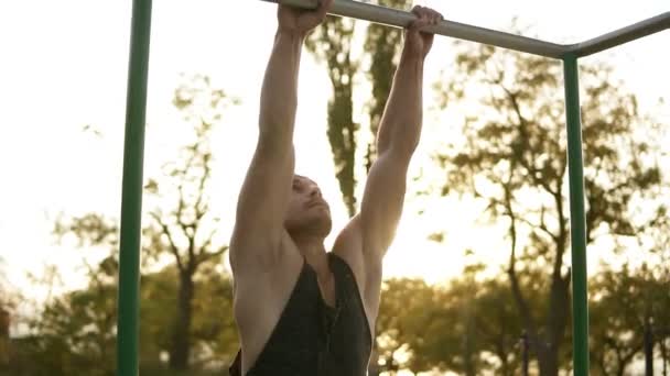 Pohledný muž s rozcvičku školení na hrazdě venku. Má gymnastické kousky na kříž v parku. Objektiv světlice na pozadí — Stock video