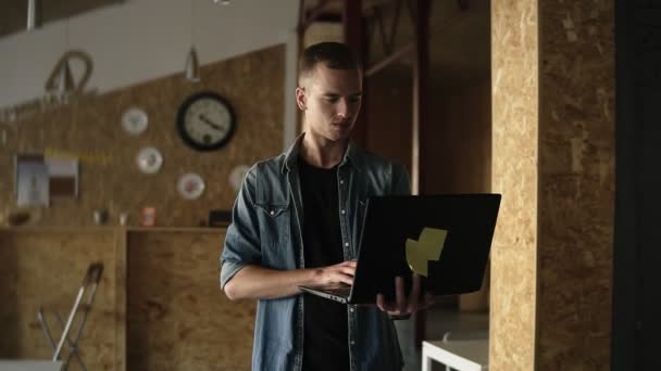 Attractive concentrada jovem homem de negócios está andando com um laptop em sua mão e digitação. Homem caucasiano trabalhando em local de trabalho público com parede de tijolo interior. Movimento lento — Vídeo de Stock