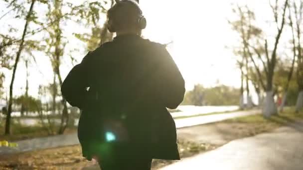 Homme paisible courir et écouter de la musique préférée à travers des écouteurs. Jogging dans le parc vert du matin en vêtements décontractés noirs. Au ralenti. Vue rare — Video