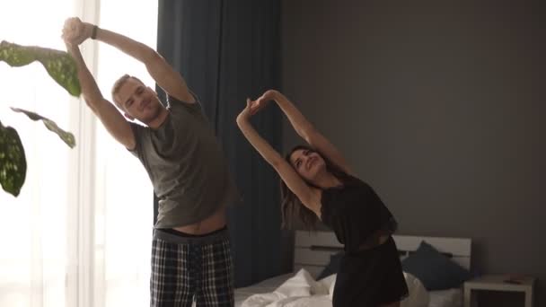 Кавказький пара робить вправу біля ліжка, після того, як вони прокинулися вранці. Молоді пара робити вправи на розтяжку і цілуватися. Вид спереду — стокове відео