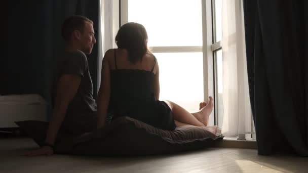 Pareja romántica en pijama sentada en el suelo por la mañana. Hermosa joven mujer caucásica adulta y hombre sentado en frente de la ventana a la luz del sol derramado en la habitación y charlando. Vista rara — Vídeo de stock